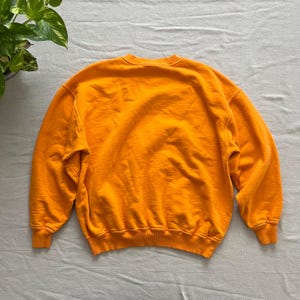 May include: A bright orange sweatshirt with a crew neck and long sleeves. The garment is made of a soft, textured fabric. The sweatshirt is laid flat on a light gray surface. The back of the sweatshirt is visible.