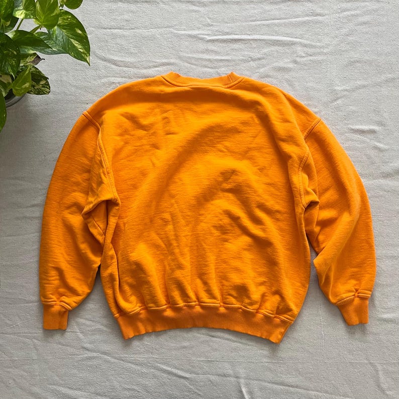 May include: A bright orange sweatshirt with a crew neck and long sleeves. The garment is made of a soft, textured fabric. The sweatshirt is laid flat on a light gray surface. The back of the sweatshirt is visible.