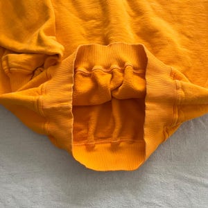 May include: Close-up of a mustard yellow sweatshirt. The image highlights the ribbed neckline, cuffs, and hem. The interior of the sweatshirt is a soft, fleecy material. The garment appears to be made of a thick, warm fabric.