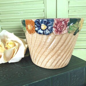 May include: A ceramic planter with a beige, crackled glaze finish and a diagonal ribbed texture. The rim is decorated with a row of three-dimensional, hand-painted flowers in shades of orange, blue, pink, and green, encircling the top edge.