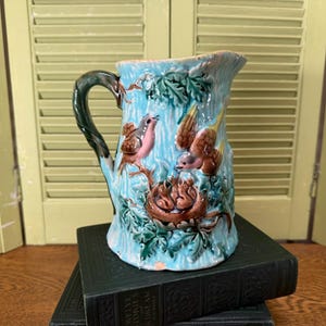 May include: A light blue ceramic pitcher featuring a raised, three-dimensional design of brown birds perched on branches and a nest with small hatchlings. The handle is dark green, and the surface is decorated with green leaves and textured details.