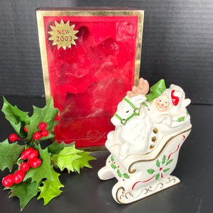 May include: A white ceramic Santa Claus in a sleigh figurine with gold accents, holly and berry details, and a green reindeer. The figurine is next to a red velvet-lined box with "NEW 2003" text. A sprig of holly and berries is in the foreground.
