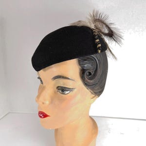 May include: A black beret-style hat with a decorative feather accent. The hat is made of a dark, possibly felt-like material. The feathers are a mix of light brown and gray. The hat is displayed on a mannequin head.