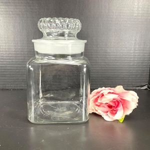 May include: A clear, square glass apothecary jar with a decorative, textured lid. The jar has a white rim and is next to a pink and red flower. The jar is empty and sits on a dark surface.