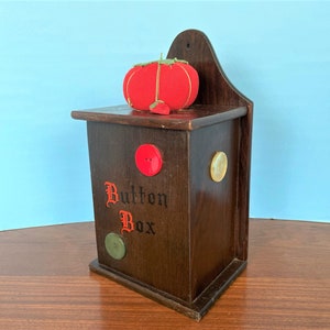 May include: A wooden button box with a red fabric pincushion on top. The box has a sign that says "Button Box" and has three buttons on the front.