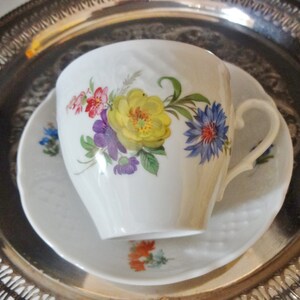 May include: A white porcelain teacup and saucer with a floral design. The cup has a heart-shaped handle and the saucer has a scalloped edge. The flowers are painted in shades of red, yellow, blue, and purple.