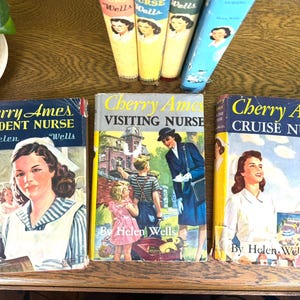 May include: A collection of vintage "Cherry Ames" novels by Helen Wells. The books feature colorful illustrations and titles like "Student Nurse," "Visiting Nurse," and "Cruise Nurse." The books are displayed on a wooden surface.