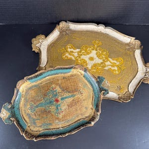 May include: Two decorative trays with ornate designs. The larger tray has a gold and white color scheme with a gold floral pattern. The smaller tray has a teal and brown color scheme with a teal and red floral pattern.