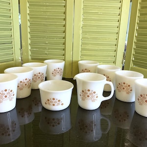 May include: Set of ten white ceramic mugs with a brown floral pattern. The mugs are arranged in a semi-circle on a black surface.