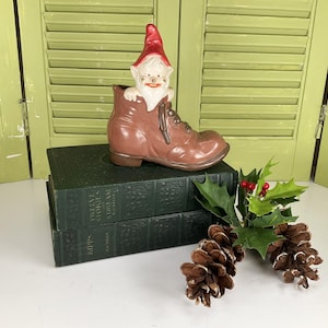 May include: A ceramic gnome figurine wearing a red hat and sitting inside a brown leather boot. The boot is sitting on top of two green hardcover books with gold lettering. The books are titled "Favorite Stories" and "Whims". There are also pine cones and holly sprigs in the image.