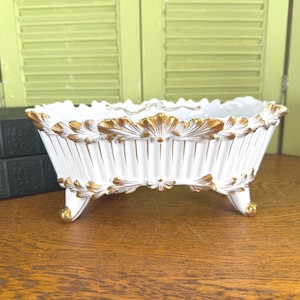 May include: A white porcelain footed bowl with gold accents. The bowl has a scalloped rim and is decorated with gold leaf designs. The bowl is on a wooden surface with green shutters in the background. The bowl is approximately 10 inches long.
