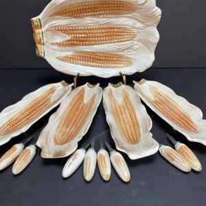 May include: A ceramic corn-shaped serving platter with matching individual corn-shaped plates and corn cob holders. The platter and plates are white with brown and yellow detailing. The corn cob holders are white with brown and yellow detailing.