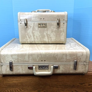 May include: Two vintage suitcases with a cream-colored, textured finish. The smaller suitcase is stacked on top of the larger suitcase. Both suitcases have gold-colored hardware and a lock.