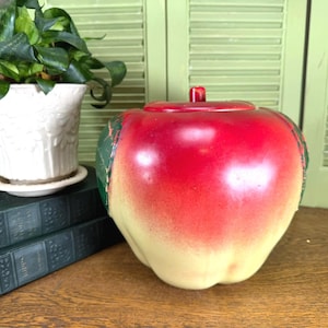 May include: A ceramic apple-shaped jar with a removable lid, featuring a gradient finish that transitions from a vibrant red top to a pale yellow base, accented with green leaf details on the sides, designed as a functional storage container.