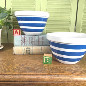 May include: Two white ceramic mixing bowls featuring horizontal blue stripes. One bowl is placed on top of two stacked vintage books, while the other sits to the right. A red block with the letter A and a green block with the letter B are present.