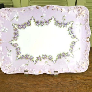 May include: A rectangular porcelain serving tray with a white center and a light lavender border. The border features a painted garland of purple and green violets, accented with raised gold-toned scrollwork and fleur-de-lis motifs.