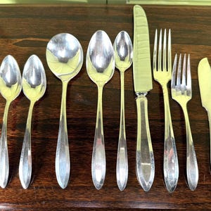 May include: A collection of silver-colored flatware, including spoons, forks, and knives, arranged on a dark wooden surface. The utensils have a simple, elegant design, reflecting a classic aesthetic. The arrangement suggests a formal dining setting.