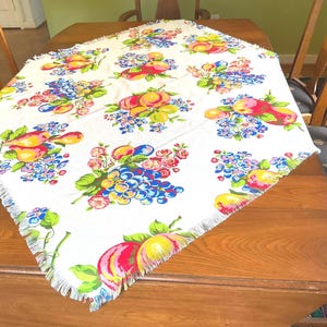 May include: White tablecloth with a colorful fruit and floral pattern. The design includes red, yellow, and purple fruits, along with blue and pink flowers. The tablecloth has a fringed edge and is placed on a wooden table.