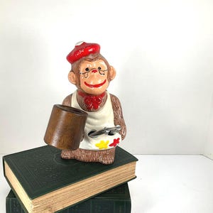May include: A vintage monkey figurine wearing a red beret, glasses, and a white apron, holding a wooden cup and a palette with paint. The monkey is standing on a stack of green books.