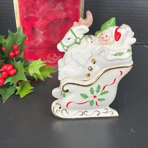 May include: A white ceramic Christmas sleigh figurine with a reindeer pulling Santa Claus. The sleigh is decorated with gold trim, holly, and red ribbons. The reindeer has brown antlers and a green harness. The figurine is likely a collectible holiday decoration.