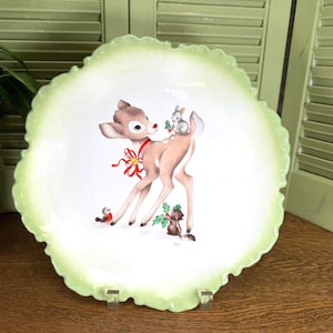 May include: A round ceramic plate with a ruffled edge and a light green border. The center features a brown fawn with a red ribbon around its neck, a small gray rabbit on its back, a brown squirrel holding holly, and a small bird.