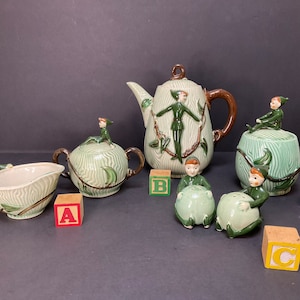 May include: A green and brown ceramic tea set with elf figurines. The set includes a teapot, creamer, sugar bowl, and two salt and pepper shakers. The set is decorated with green leaves and vines.