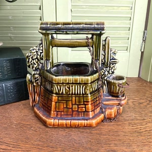May include: A ceramic wishing well planter with a brick-like design in shades of brown, green, and orange. The planter features a small bucket and chain detail. The words "WISHING WELL" are visible on the front. A decorative item.