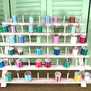 May include: A tiered wooden thread rack holding multiple spools of sewing thread in various colors including blue, green, pink, red, and white. The rack features five horizontal shelves with vertical pegs to secure each individual spool of thread.