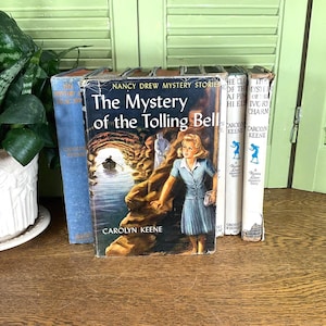 May include: A collection of vintage Nancy Drew mystery books. The central book, "The Mystery of the Tolling Bell," features a cover illustration of a young woman in a blue dress. Other books are visible, with titles partially visible.
