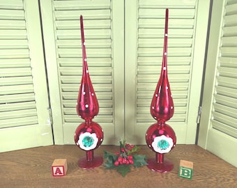 Puede incluir: Dos adornos rojos para la parte superior del árbol de Navidad con lunares blancos y una base decorativa. Cada adorno tiene una base redonda con un diseño floral blanco y verde. Los adornos se exhiben sobre una superficie de madera.