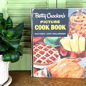 May include: Vintage Betty Crocker's Picture Cook Book. The cover features a pie, roasted chicken, and corn on the cob. The book is revised and enlarged. A green plant in a white pot is to the left of the book.