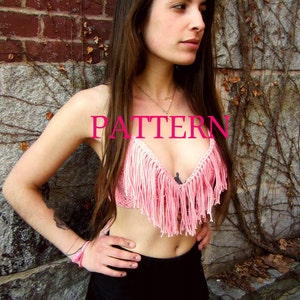May include: A pink crocheted top with fringe detailing. The top is worn by a person standing in front of a brick wall with vines.