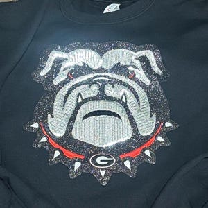 May include: Black sweatshirt featuring a glittery bulldog design. The bulldog has silver and black details, red accents, and a red collar with white spikes. The design includes the letter "G" inside a circle.