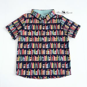 May include: Short-sleeved button-down shirt with a navy blue background and a colorful bookshelf pattern. The collar and inner lining are a light blue. The shirt has black buttons and the text "Lilies & Lizards" is visible.
