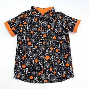 May include: Black and gray button-up shirt with a Halloween theme. The shirt features a pattern of skeletons, spiderwebs, and pumpkins. The collar and sleeve cuffs are orange. The words "BOO!" and "BAD BONE" are printed on the shirt.