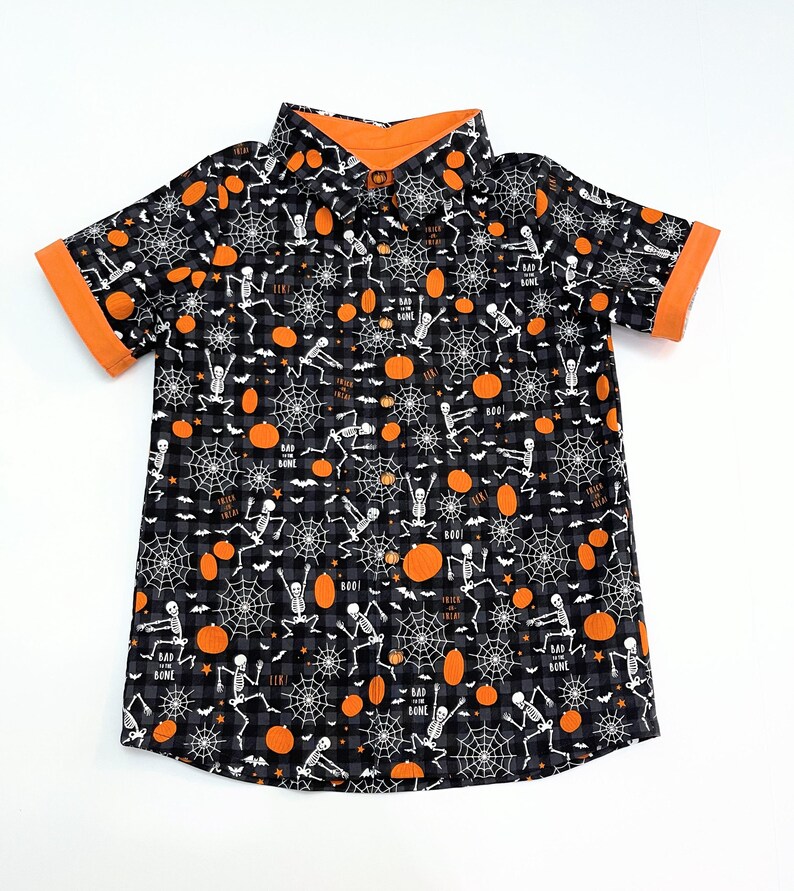 May include: Black and gray button-up shirt with a Halloween theme. The shirt features a pattern of skeletons, spiderwebs, and pumpkins. The collar and sleeve cuffs are orange. The words "BOO!" and "BAD BONE" are printed on the shirt.