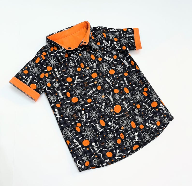 May include: A short-sleeved button-down shirt with a Halloween-themed pattern. The shirt features a black and gray checkered background with orange pumpkins, white skeletons, and spiderwebs. The collar and sleeve cuffs are orange.