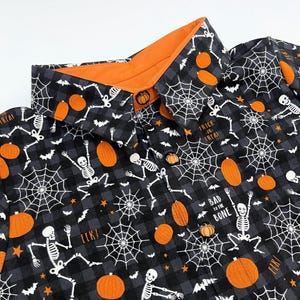 May include: Black and gray Halloween-themed button-up shirt with an orange collar. The shirt features a pattern of orange pumpkins, white spiderwebs, skeletons, and bats. The text "Trick or Treat" and "Bad to the Bone" are visible.