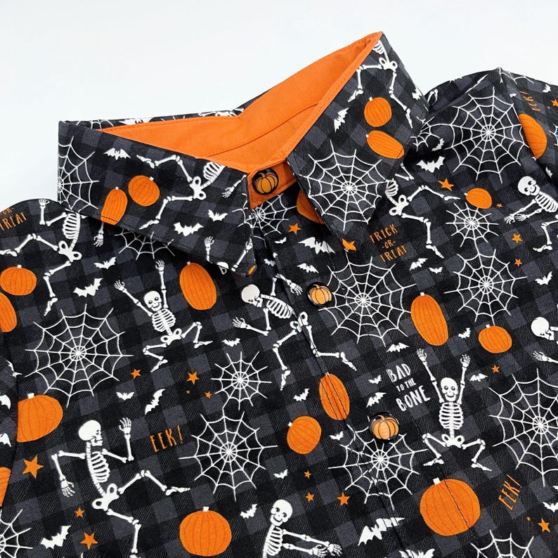 May include: Black and gray Halloween-themed button-up shirt with an orange collar. The shirt features a pattern of orange pumpkins, white spiderwebs, skeletons, and bats. The text "Trick or Treat" and "Bad to the Bone" are visible.