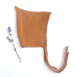 May include: A pointed bonnet in a solid, warm ochre-brown color with a subtle herringbone weave pattern. The headwear features two long, matching fabric ties extending from the base and is displayed alongside a single sprig of purple lavender.