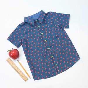 May include: A short-sleeved button-down shirt in a dark blue fabric featuring a repeating pattern of small red apples. The shirt has a pointed collar, a single chest pocket, and bright green buttons. A red apple and a yellow pencil are nearby.