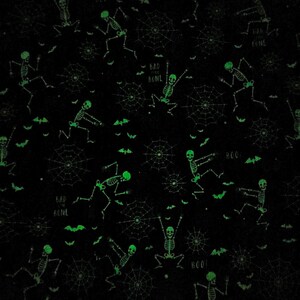 May include: Black fabric with a repeating pattern of glowing green skeletons in various poses, spiderwebs, and bat silhouettes. The words "BAD BONE" and "BOO!" are also scattered across the design. This Halloween-themed fabric is suitable for various craft projects.