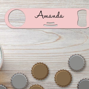 Puede incluir: Abrebotellas de metal rosa con el nombre "Amanda" grabado en él. El abrebotellas está rodeado de tapas de botellas sobre una superficie de madera.