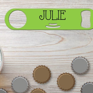 Puede incluir: Abrebotellas verde con el nombre "JULIE" impreso en él. El abrebotellas está sobre una superficie de madera con varias tapas de botellas esparcidas a su alrededor.