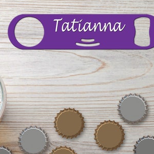 Puede incluir: Abrebotellas de metal morado con el nombre "Tatianna" grabado. El abrebotellas está sobre una superficie de madera clara con varias tapas de botellas.