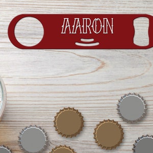 Puede incluir: Un abridor de botellas rojo con el nombre "AARON" impreso en él. El abridor está sobre una superficie de madera con varias tapas de botellas esparcidas a su alrededor.