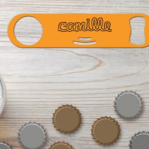 Puede incluir: Abrebotellas de metal naranja con el nombre "Camille" grabado en él. El abrebotellas está sobre una superficie de madera clara con varias tapas de botellas esparcidas a su alrededor.