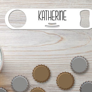 Puede incluir: Un abridor de botellas de metal blanco con el nombre "Katherine" impreso en él. El abridor de botellas está sobre una superficie de madera con varias tapas de botellas esparcidas a su alrededor.
