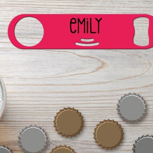 Puede incluir: Un abridor de botellas de metal rosa con el nombre "EMILY" impreso en él. El abridor está sobre una superficie de madera clara con varias tapas de botellas esparcidas a su alrededor.