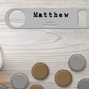 Puede incluir: Un abridor de botellas plateado con el nombre "Matthew" grabado en él. El abridor está rodeado de tapas de botellas sobre una superficie de madera.